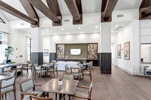 Spacious modern gathering area with wooden beams, tables, chairs, and a TV on the wall. Bright and inviting atmosphere.