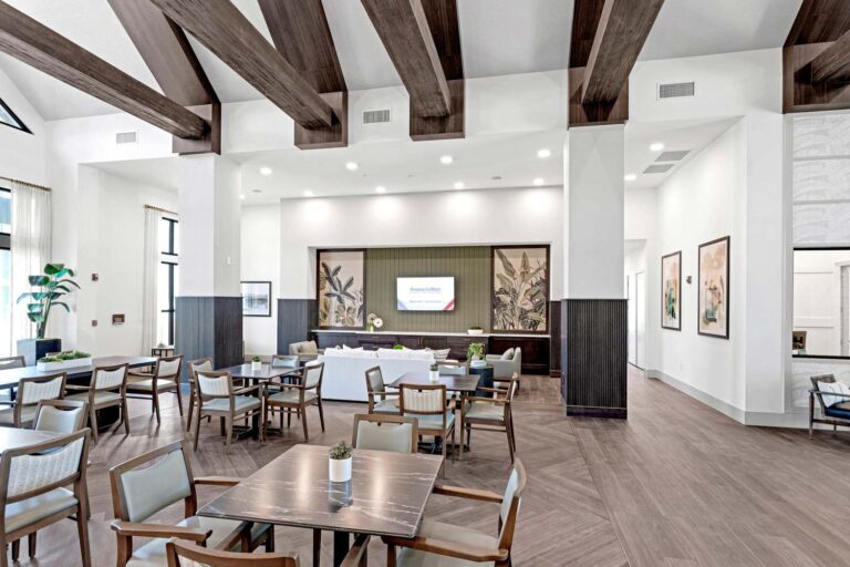 Spacious modern gathering area with wooden beams, tables, chairs, and a TV on the wall. Bright and inviting atmosphere.
