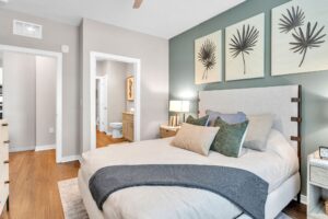 Modern bedroom with neutral tones, cozy bedding, and palm-themed art, leading to a bathroom with wood flooring.