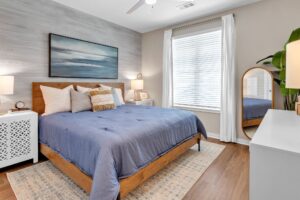 Modern bedroom with blue bedding, wooden bed frame, and decorative pillows, lit by natural light from a large window.