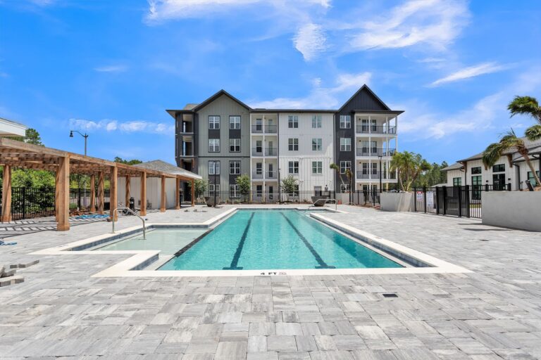 Modern apartment complex with outdoor swimming pool and lounge area under a clear blue sky.