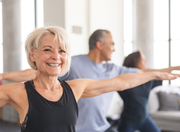 Elderly woman smiling during a yoga class, stretching arms in a bright room with other participants in the background.
